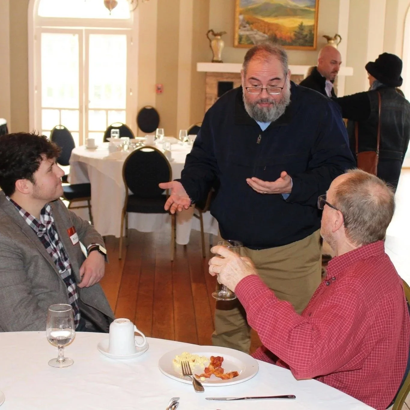 Three men are in a dining room, engaged in conversation. One man stands and gestures while the others sit at a table with partially eaten pizza. The man standing has glasses, a beard, and wears a dark jacket. The seated man to the right wears glasses and a red shirt. The other seated man has dark hair and a checked shirt. The background features tables set with glassware and a painting on the wall.
