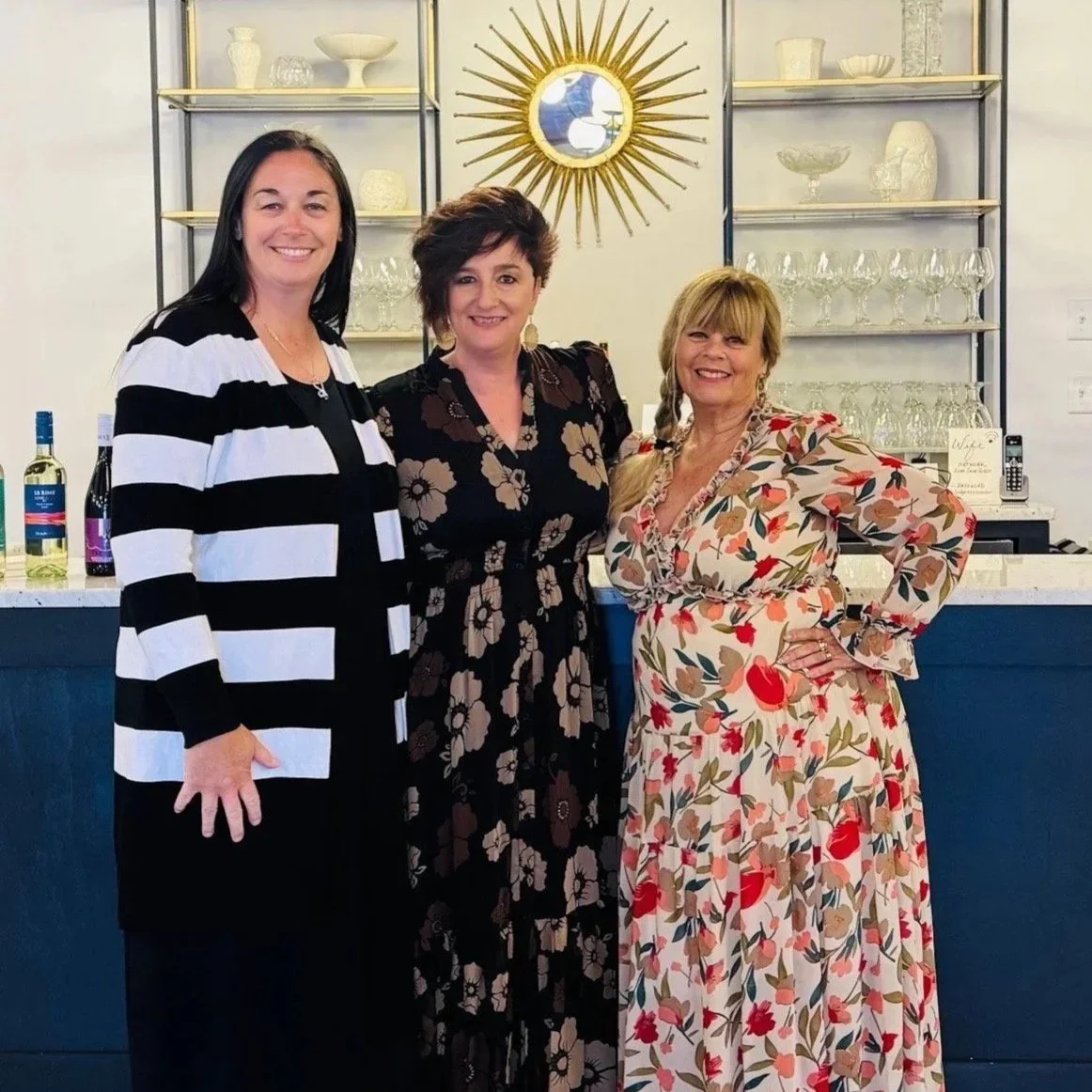 Three women dressed in stylish outfits standing together inside a bar or restaurant, smiling at the camera. Behind them are shelves with glassware and decorative items, a round gold mirror, and bottles on the counter.