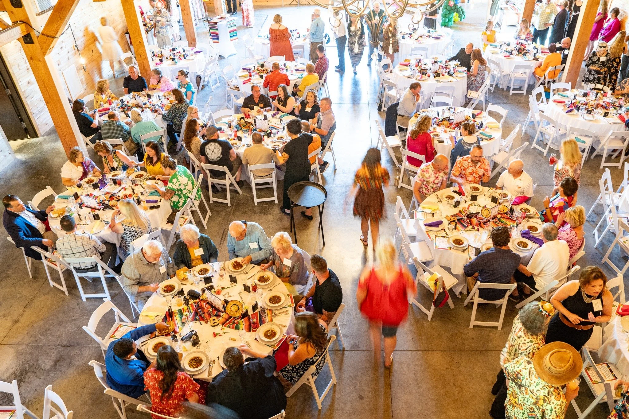 Large indoor gathering of people at round tables set for a meal, with some standing and talking, in a rustic banquet hall with wooden beams and walls.