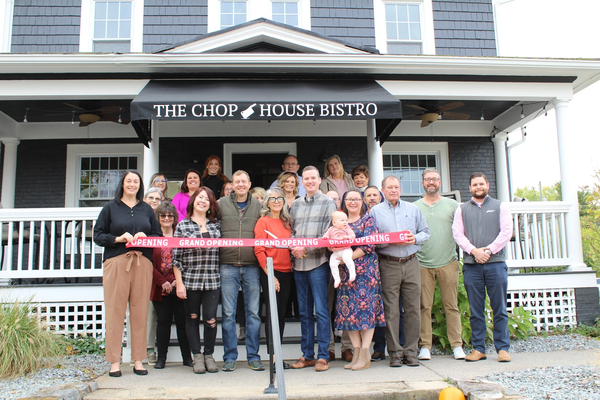 Group of people gathered in front of a house for a grand opening celebration, holding a ribbon with 'Grand Opening' written on it.