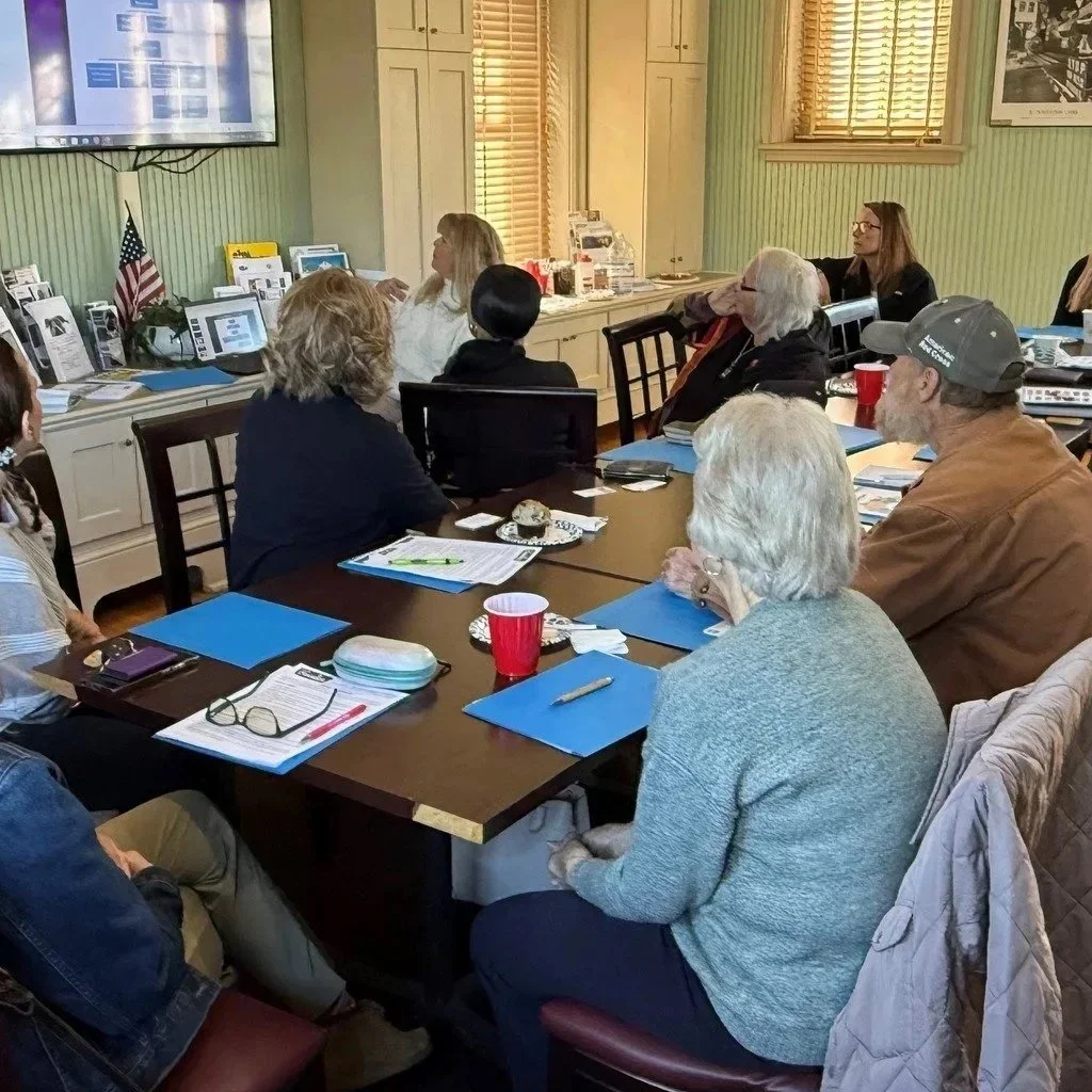 A group of people attending a meeting in a room with beige walls and wooden blinds, watching a presentation on a large screen. The group includes both men and women, some with notebooks and documents in front of them.