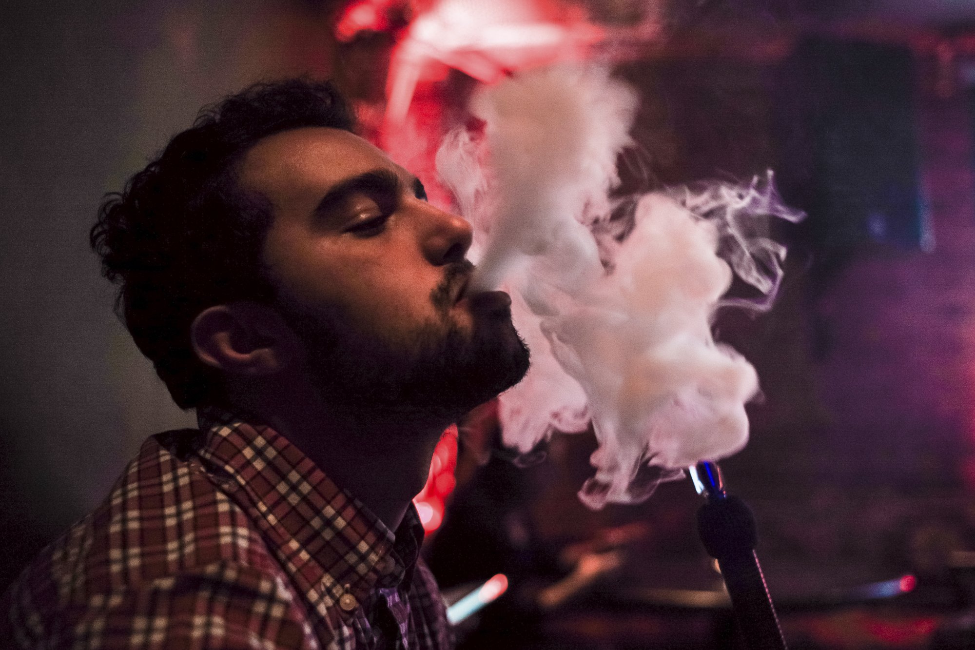 A man exhales a plume of smoke in a dimly lit hookah lounge in New York City. The soft red glow of the room and the drifting vapor create an intimate, cinematic moment—one of quiet ritual in the middle of the city’s constant motion.