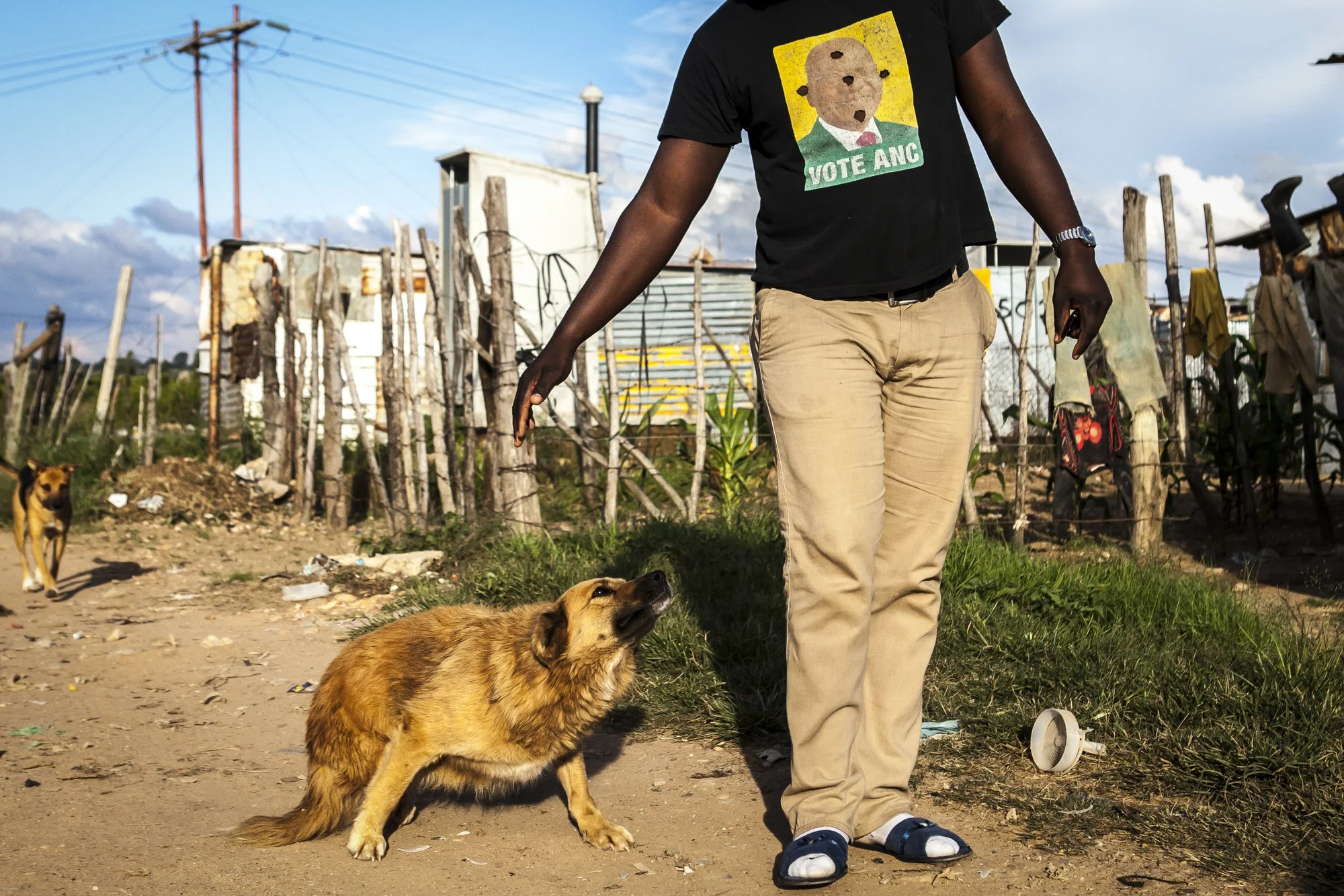 A resident walks through the informal settlement that has grown up around the abandoned Durban Deep mine, his shirt bearing the face of an ANC politician punctured by mysterious bullet-like holes — a stark, improvised commentary on the political disi