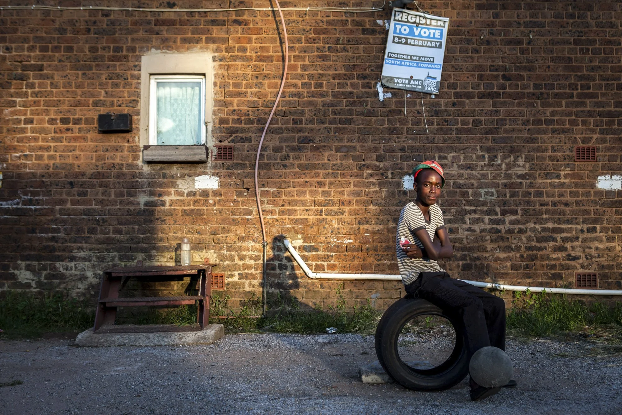 A teenaged boy sits on a tire and taps a soccer ball against his feet outside one of Durban Deep’s old hostels on the outskirts of Johannesburg, South Africa. Above him, a weathered ANC voter-registration poster hangs from the brick wall—an echo of t