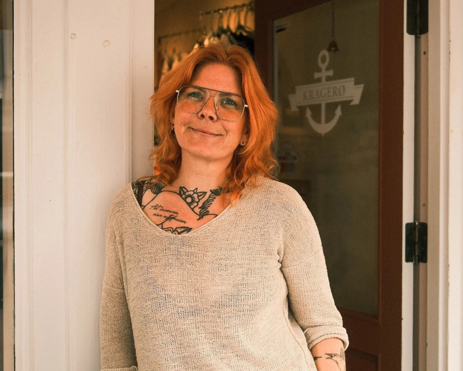 Red-haired woman wearing glasses and a beige sweater, standing near a doorway with a smile.