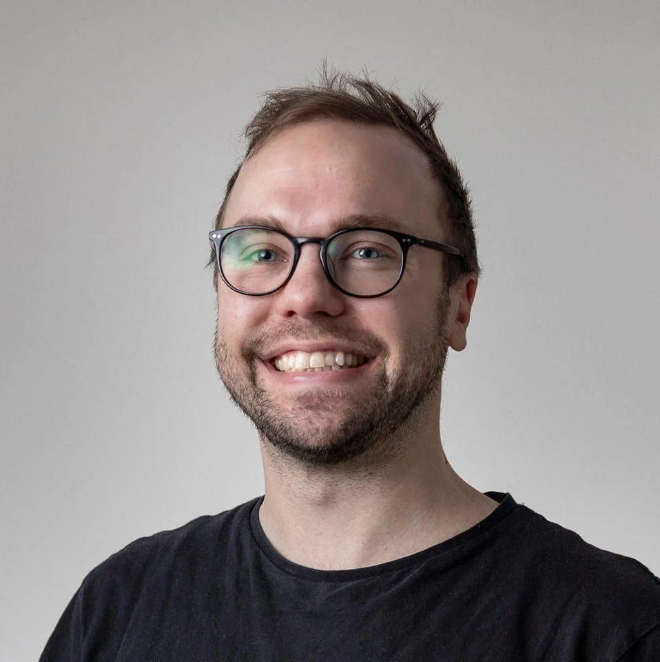Portrait of a smiling man with glasses and short brown hair, wearing a black T-shirt, against a light gray background.