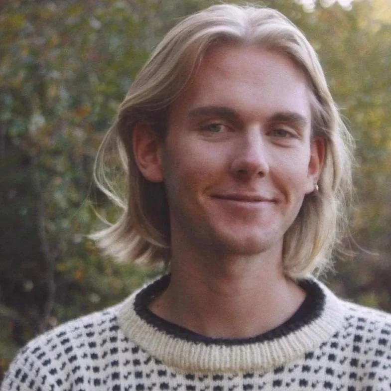 A young man with shoulder-length blonde hair smiling outdoors with trees in the background.