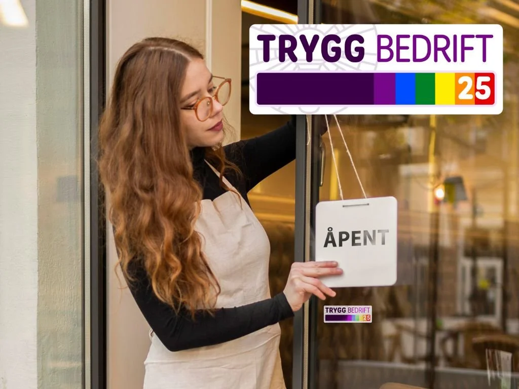 A woman with long red hair and glasses opening a door with an sign that says "OPEN." There are signs outside that say "TRYGG BEDRIFT".