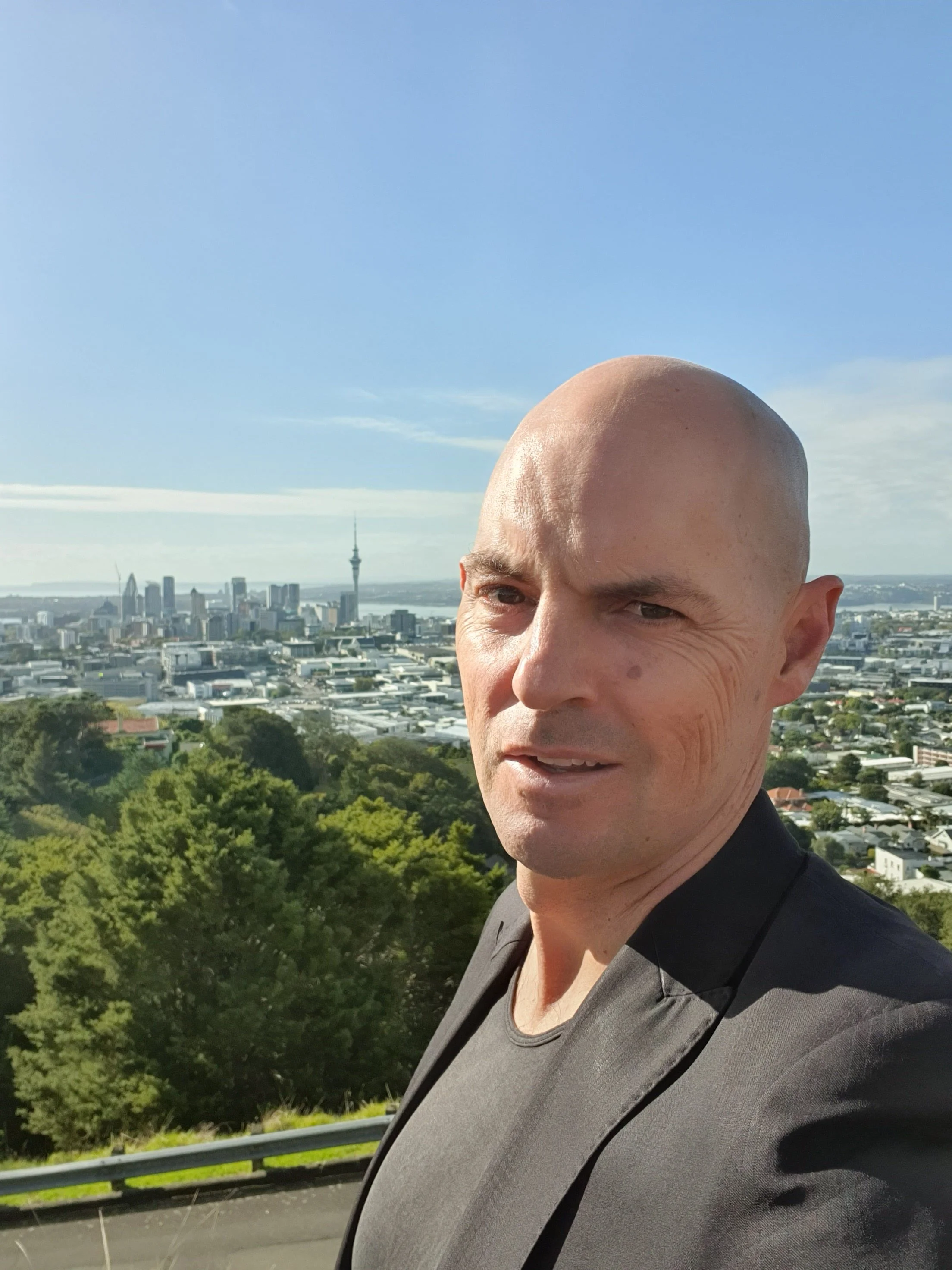Michael Sharpe in a gray blazer taking a selfie with an urban city skyline and the Sky Tower in the background under a clear blue sky.