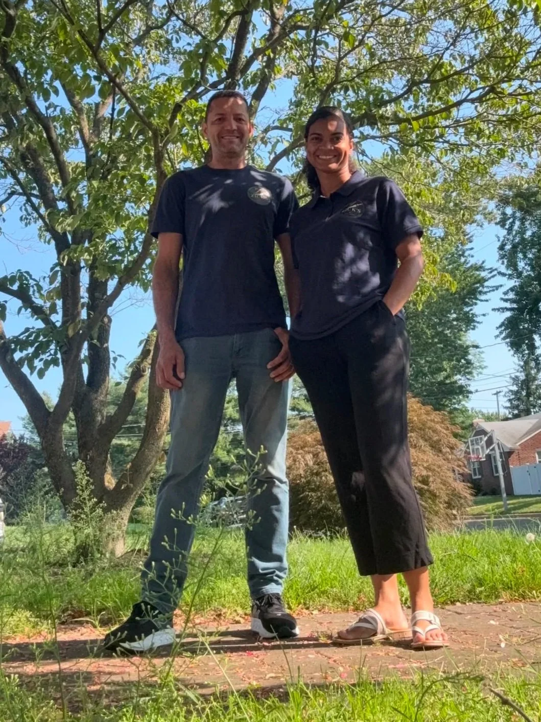 A man and woman standing outdoors on a sunny day, smiling, with green trees and a blue sky in the background, both dressed in black shirts and casual pants.