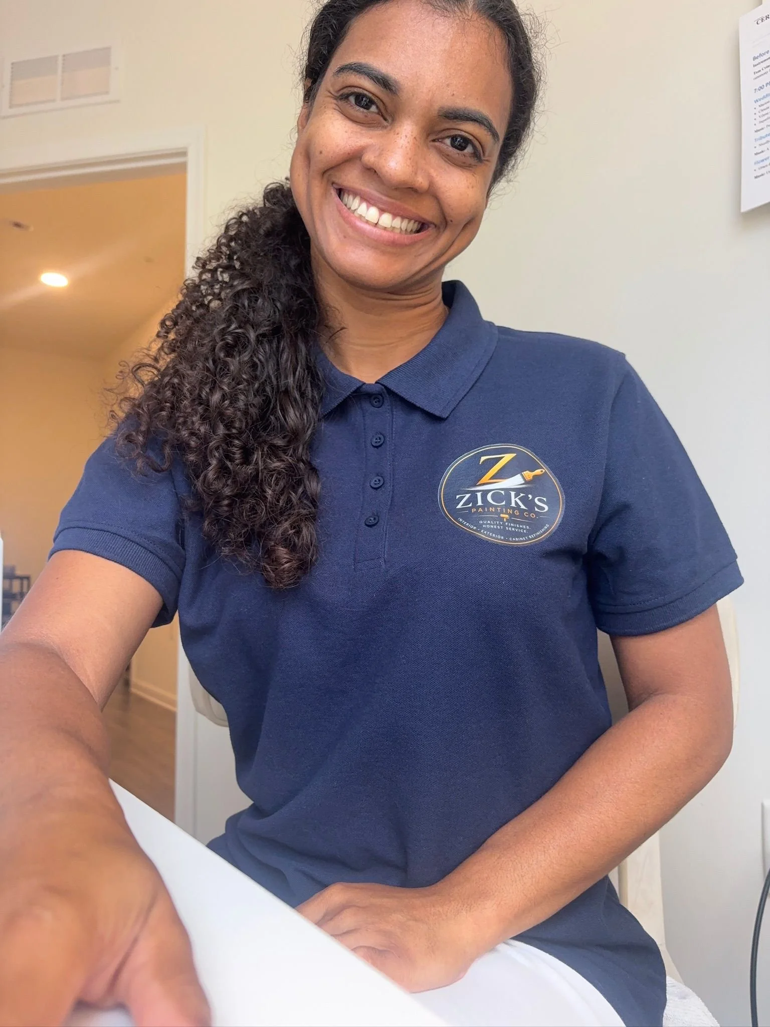 A woman smiling in a blue polo shirt with a Zick's Painting Co logo, sitting at a desk with a white surface, back against a white wall with some posters.