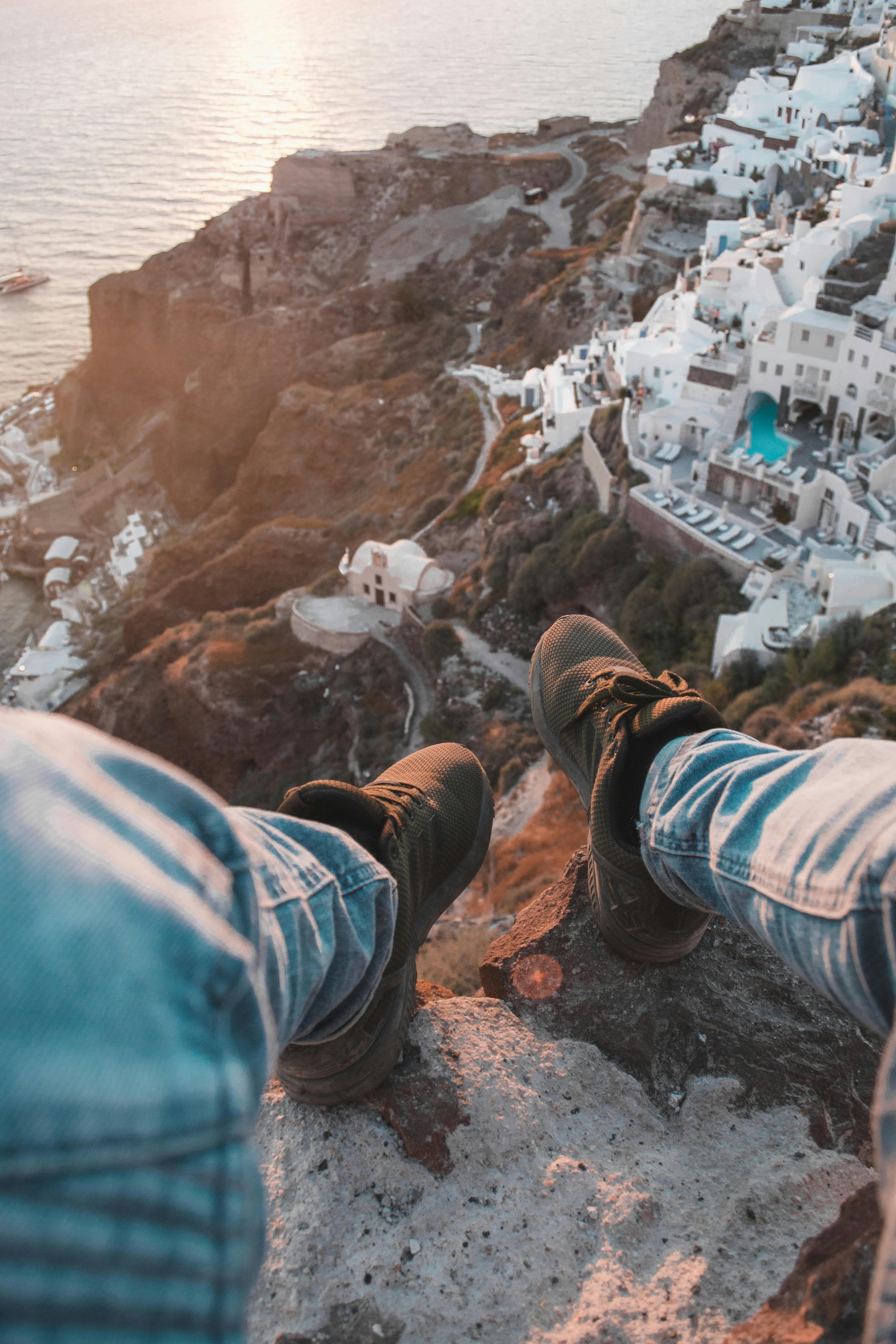 A person sitting on a cliff edge overlooking the scenic coastal town of Santorini, Greece, during sunset.