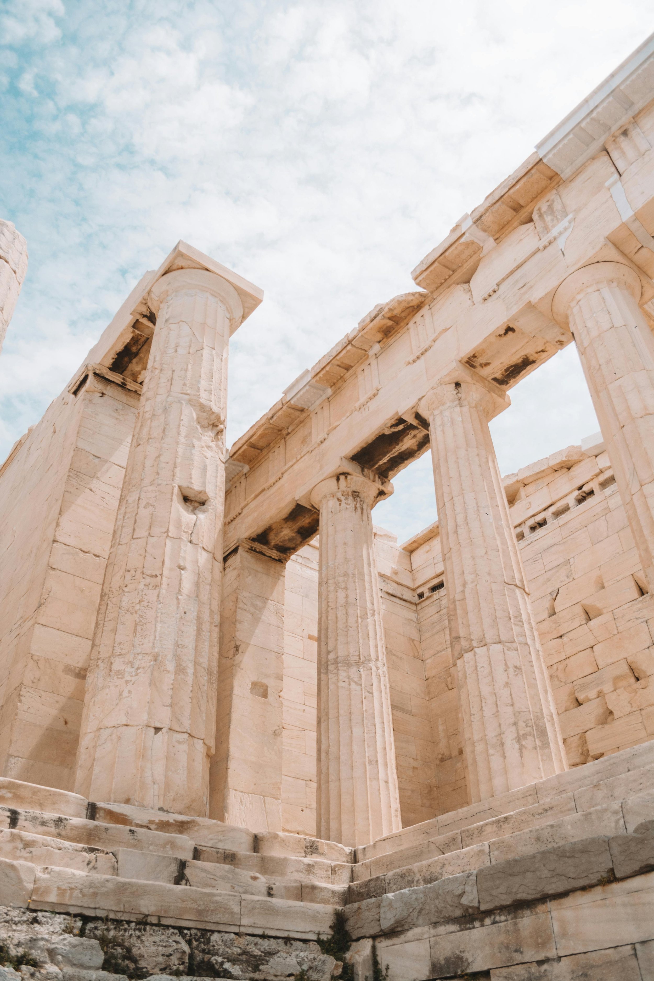 Ancient Greek ruins of a marble temple with tall, fluted columns and steps leading up to the structure, under a partly cloudy sky.
