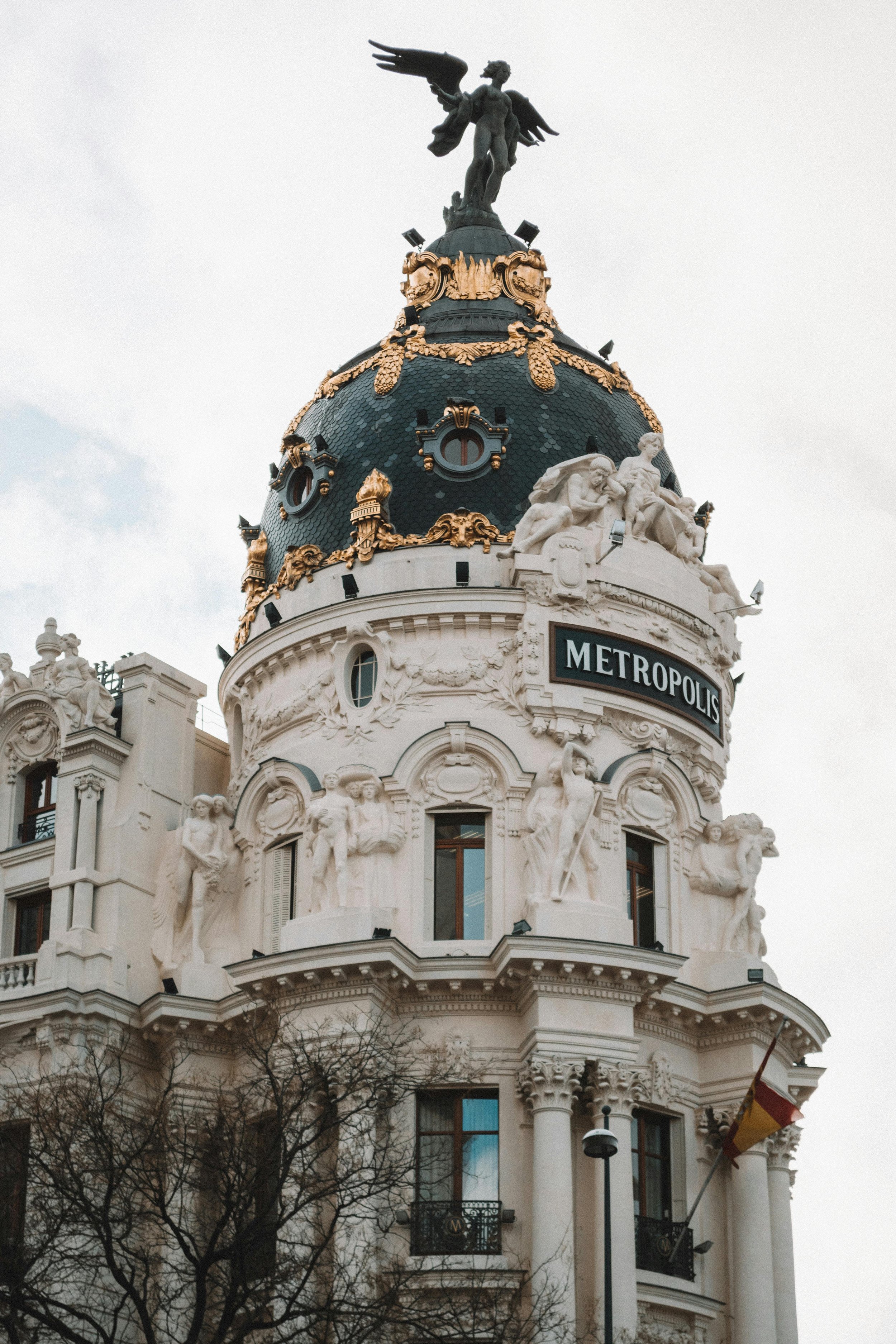 The Metropolis building in Madrid, Spain, with ornate classical architecture, sculptures, and a dome topped with a statue of a winged figure.