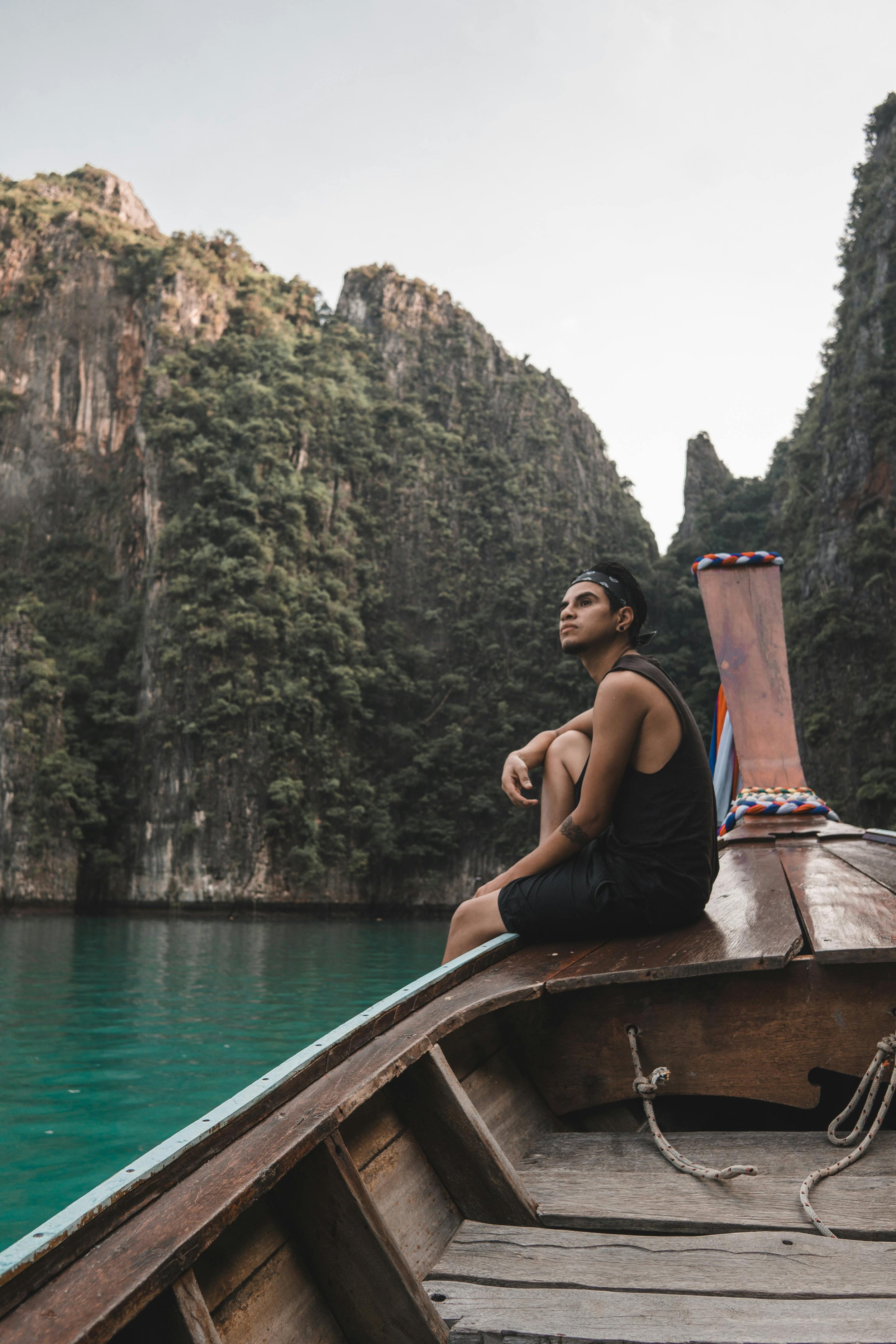 A man sits on the front of a wooden boat in a river surrounded by tall, green, rocky cliffs.