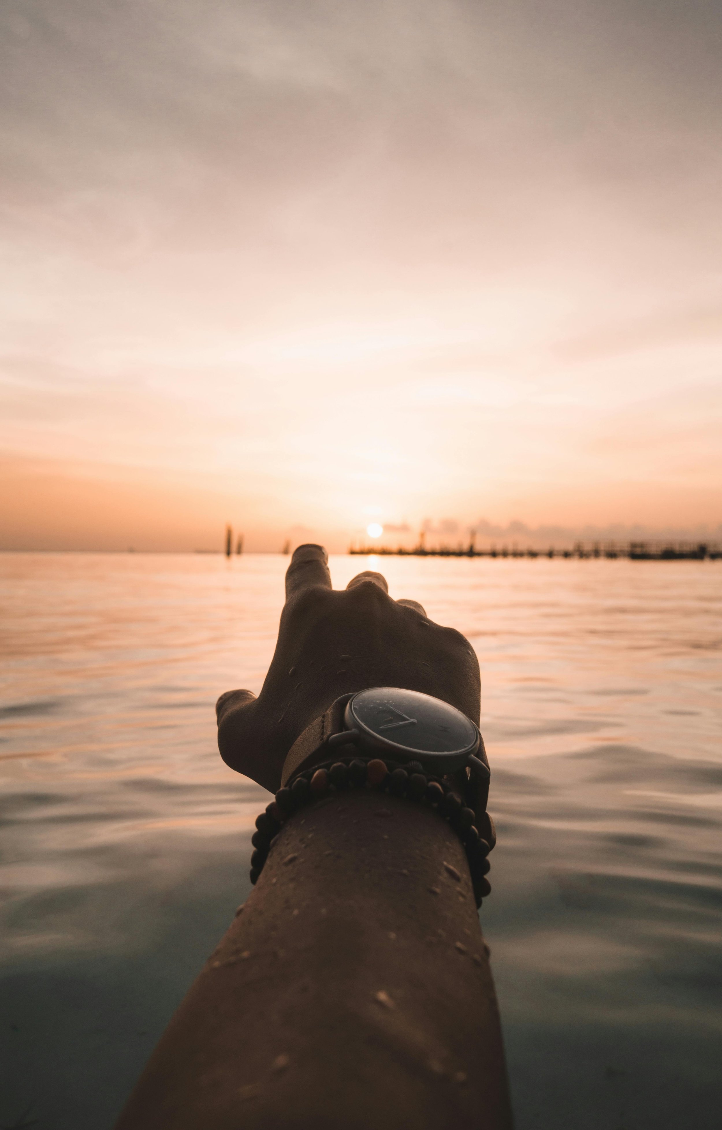 A person's arm and hand reaching out over water towards a setting sun on the horizon, wearing a watch and beaded bracelet.