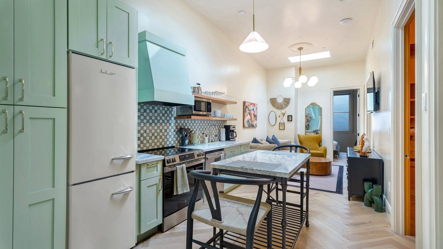 Open-concept kitchen with pastel green cabinets, marble countertop island, modern appliances, and a view into the living room with yellow and gray furniture and wall art.