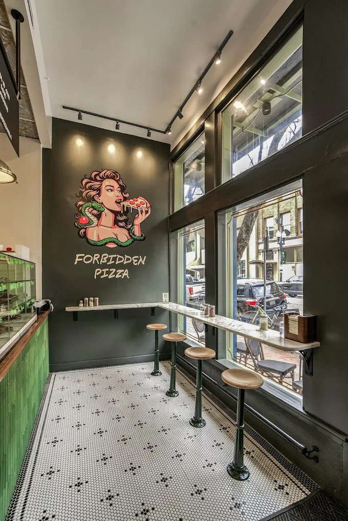 Inside a pizza restaurant with a large window, wooden stools, a dark wall with a mural of a woman eating pizza, and the text "Forbidden Pizza."