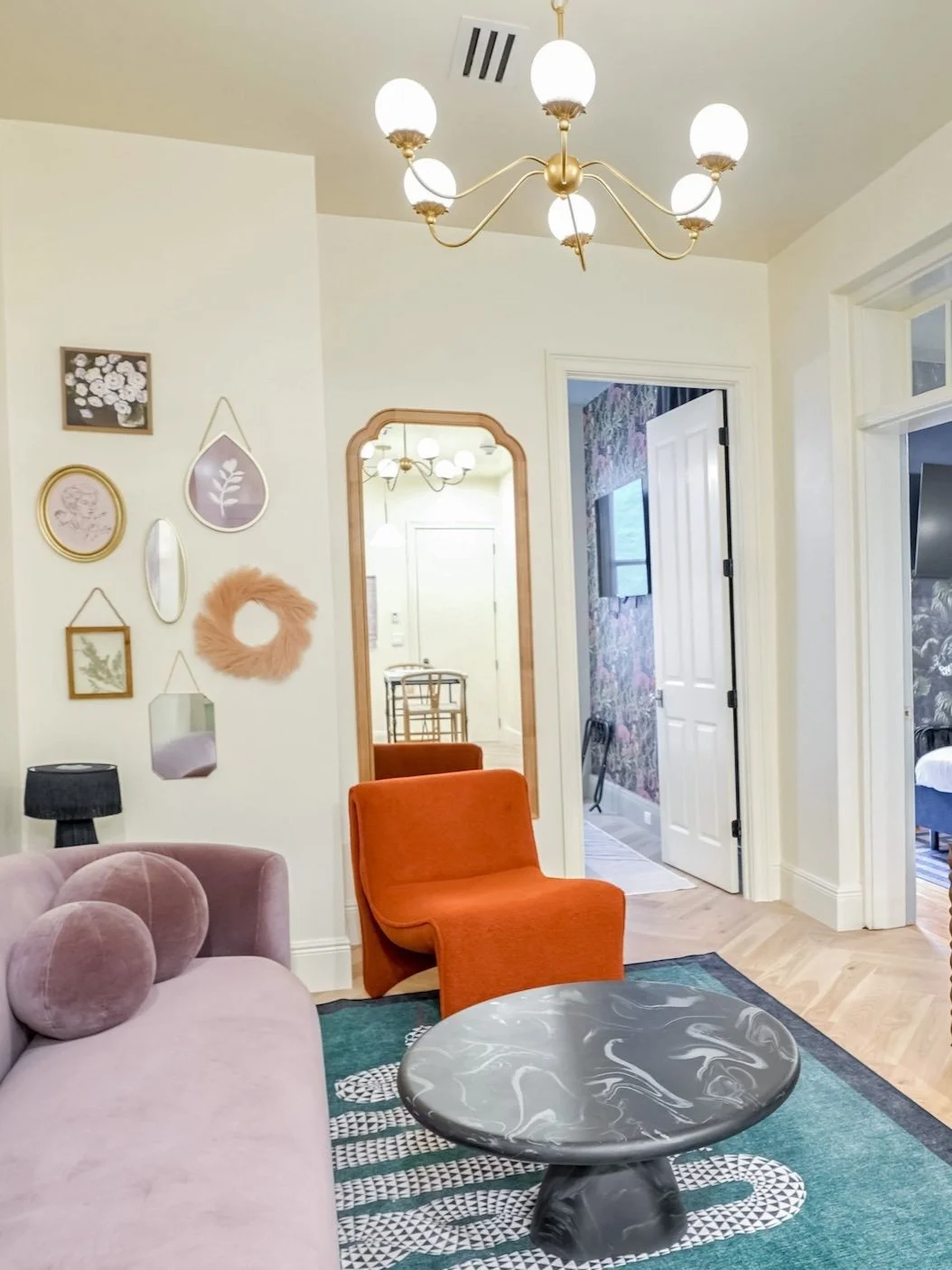 Living room with a pink velvet sofa, an orange armchair, a black side table with lamp, a marble-patterned coffee table, a chandelier, wall art, and a mirror reflecting the room. An open door leads to another room with a TV and floral wallpaper.
