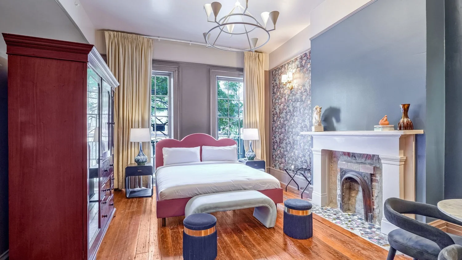 A bedroom with a pink upholstered bed, two nightstands with lamps, a large red armoire, a vintage fireplace with decorative items on the mantel, two windows with cream curtains, a modern chandelier, hardwood flooring, and an accent wall with floral wallpaper.