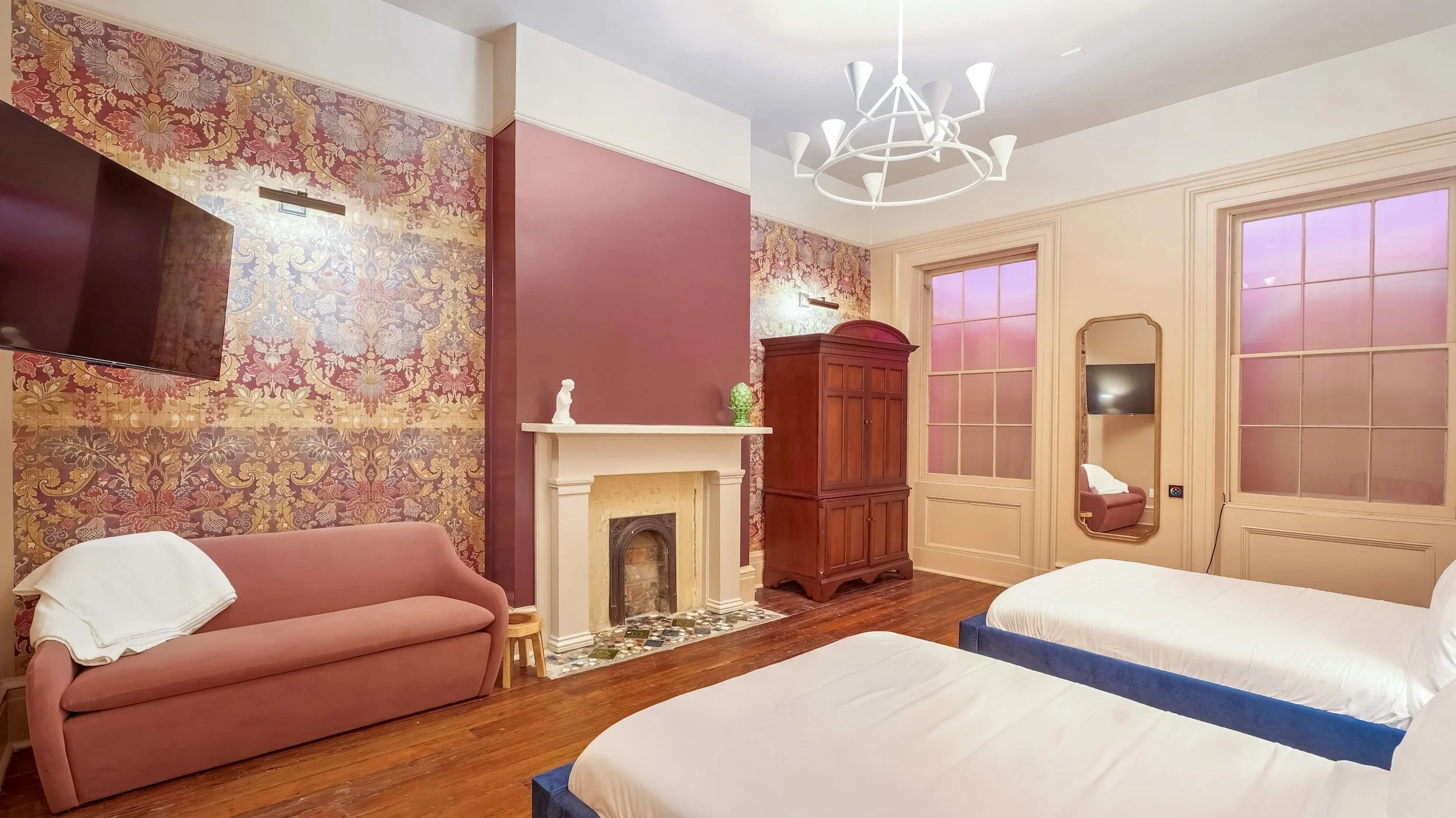 A cozy bedroom with two beds, a pink sofa with a white blanket, a white fireplace with decorative objects, wooden flooring, patterned wallpaper, and large windows with frosted pink glass.