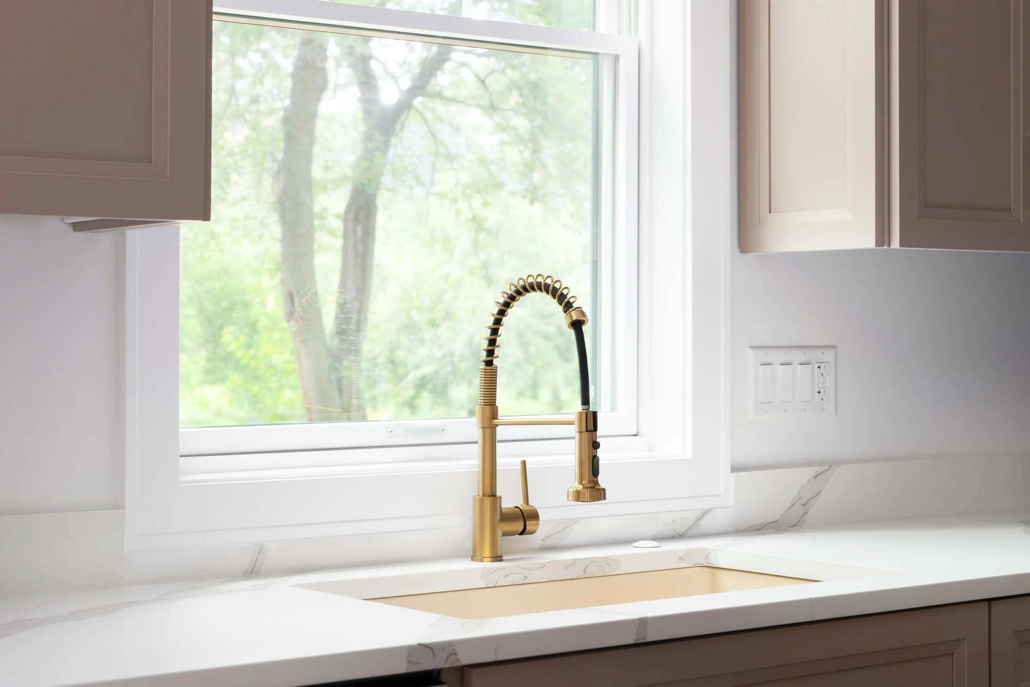 A kitchen window with a view of trees outside, a white marble countertop, a gold kitchen faucet, and beige cabinets.