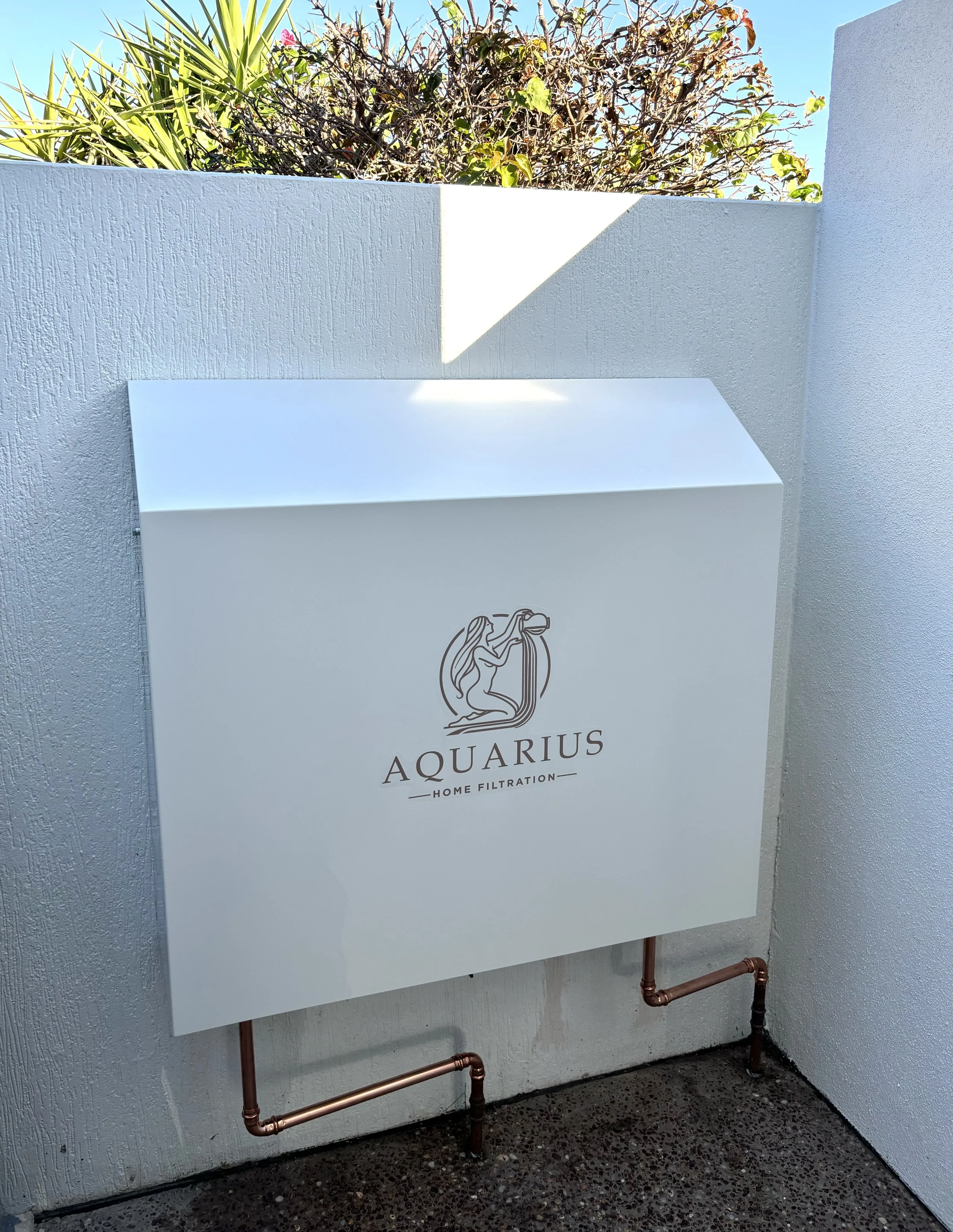 Surfmist Colourbond Whole House Filtration System featuring the Aquarius Home Filtration logo, with copper piping beneath it and a textured wall behind, and plants visible at the top.
