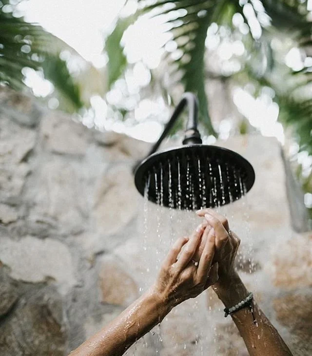 Person rinsing their hands under an outdoor shower with a stone wall and green foliage in the background.