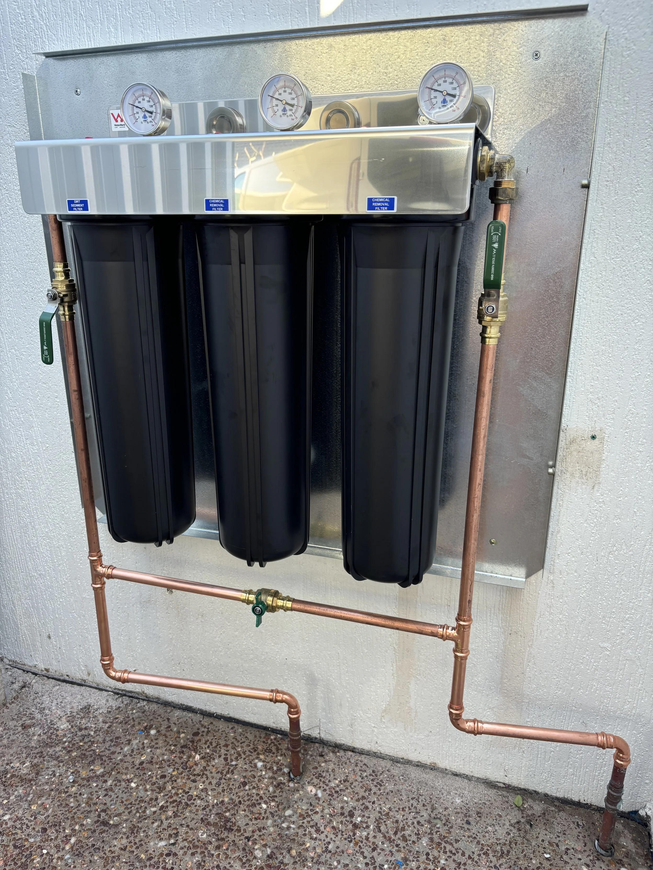 A water filtration system with three black filter cartridges mounted on a wall, copper pipes, and pressure gauges on top.