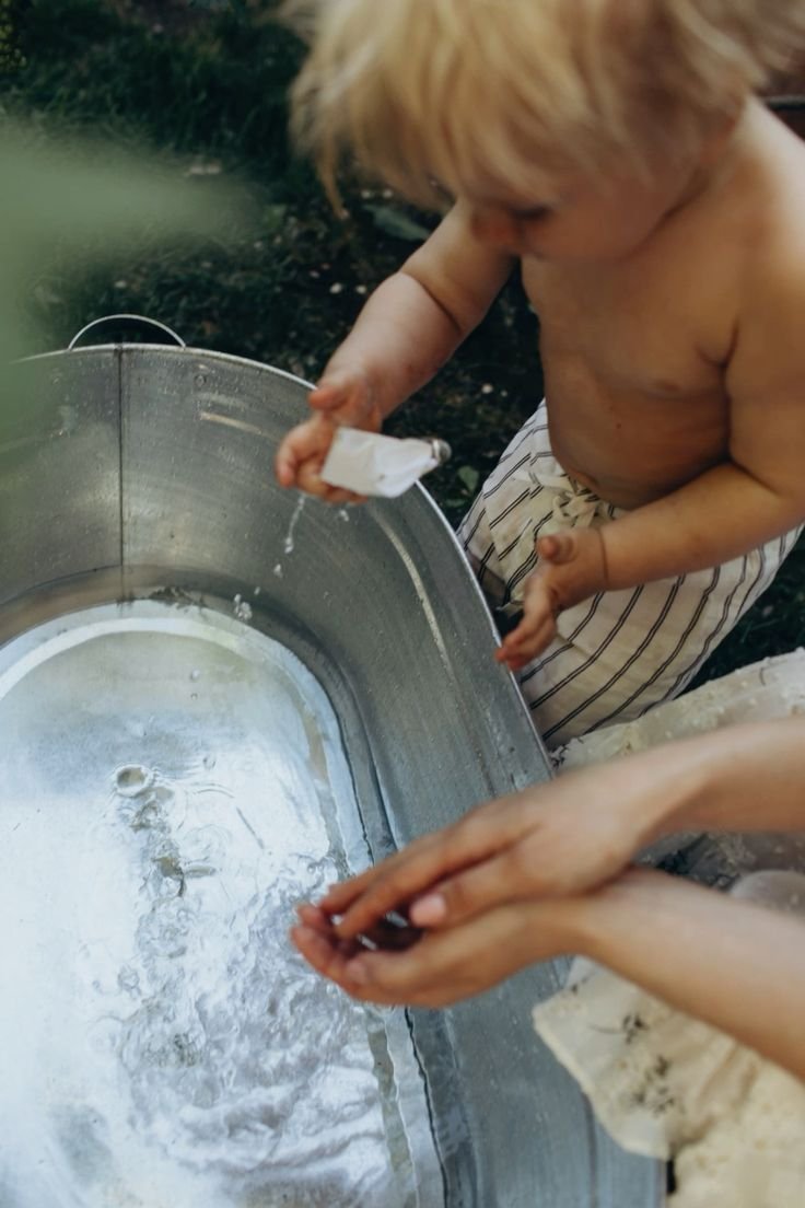 A young shirtless boy with blond hair and striped shorts is splashng water into a large metal basin with a bag. A person nearby is cupping their hands under the water. They are outdoors using the garden hose.