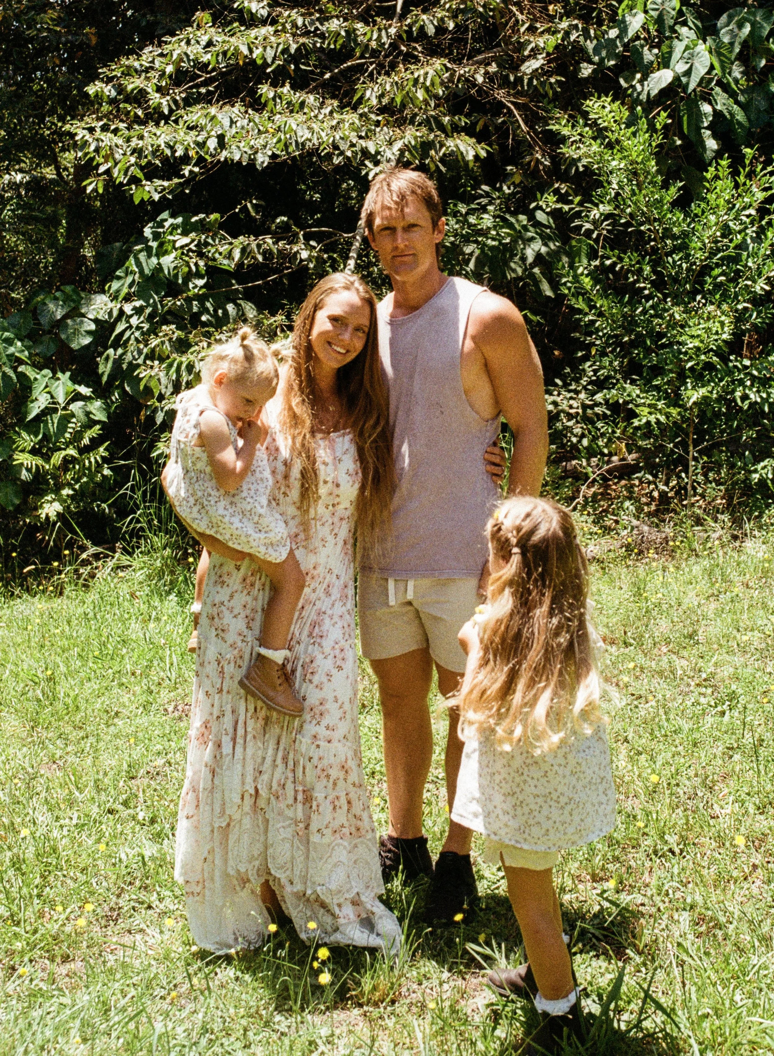 A family of four outdoors in a grassy area with trees in the background. The mother, with long hair, is wearing a white floral dress and holding a young girl with blonde hair in a white dress. The father, with short brown hair, is wearing a sleeveless shirt and shorts. A young girl with long hair and a white dress with yellow flowers is standing in front of the family, facing them.
