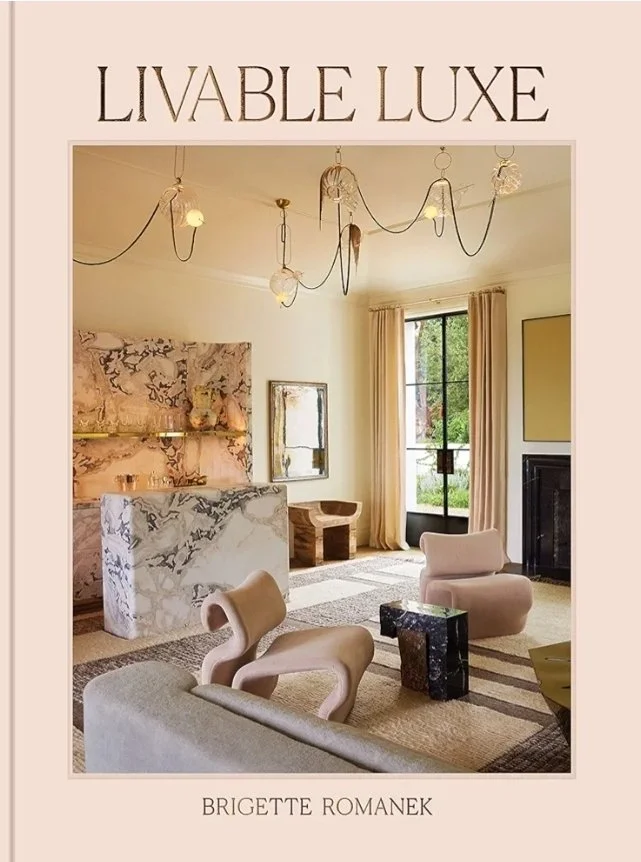 Cover of the interior design book Livable Luxe by Brigette Romanek, featuring a softly layered living space with sculptural furniture, marble surfaces, and warm neutral tones