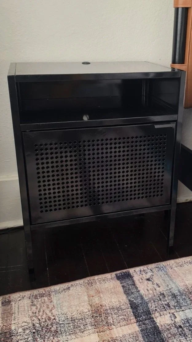 Black metal nightstand with perforated cabinet door beside the cognac upholstered bed