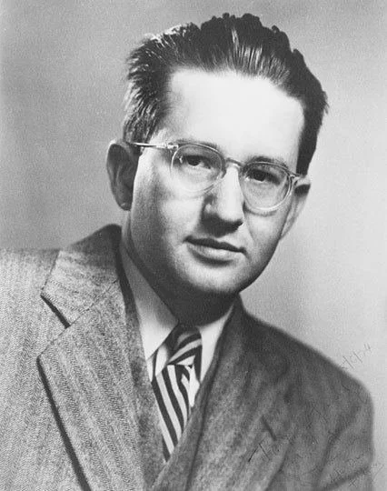 Black and white portrait of Andrew J. Galambos wearing glasses and a suit, with a striped tie.