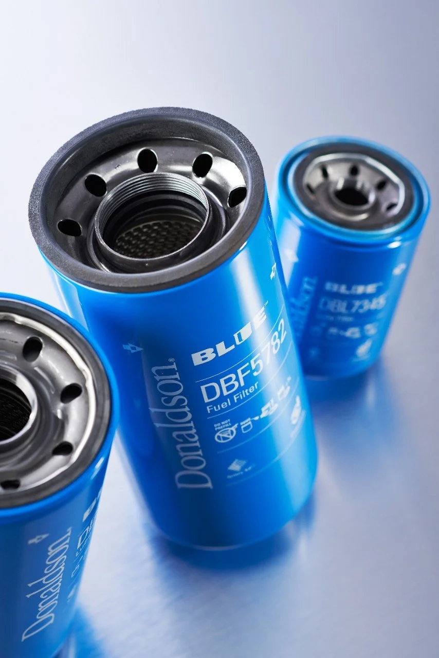 Three blue Donaldson fuel filters with metal tops, positioned on a light blue surface.