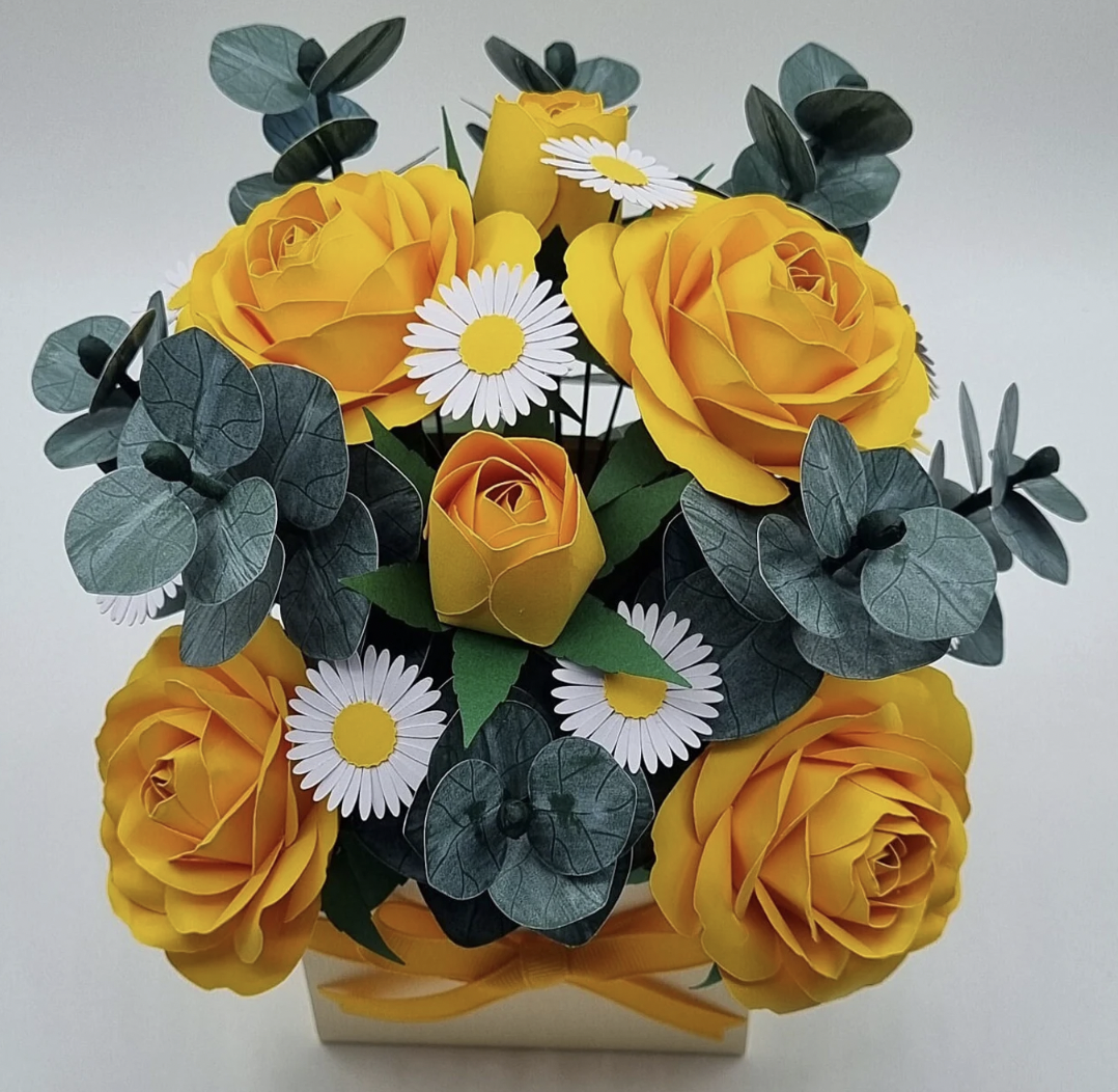 A bouquet of yellow roses, white daisies with yellow centers, and green leaves in a white square pot.