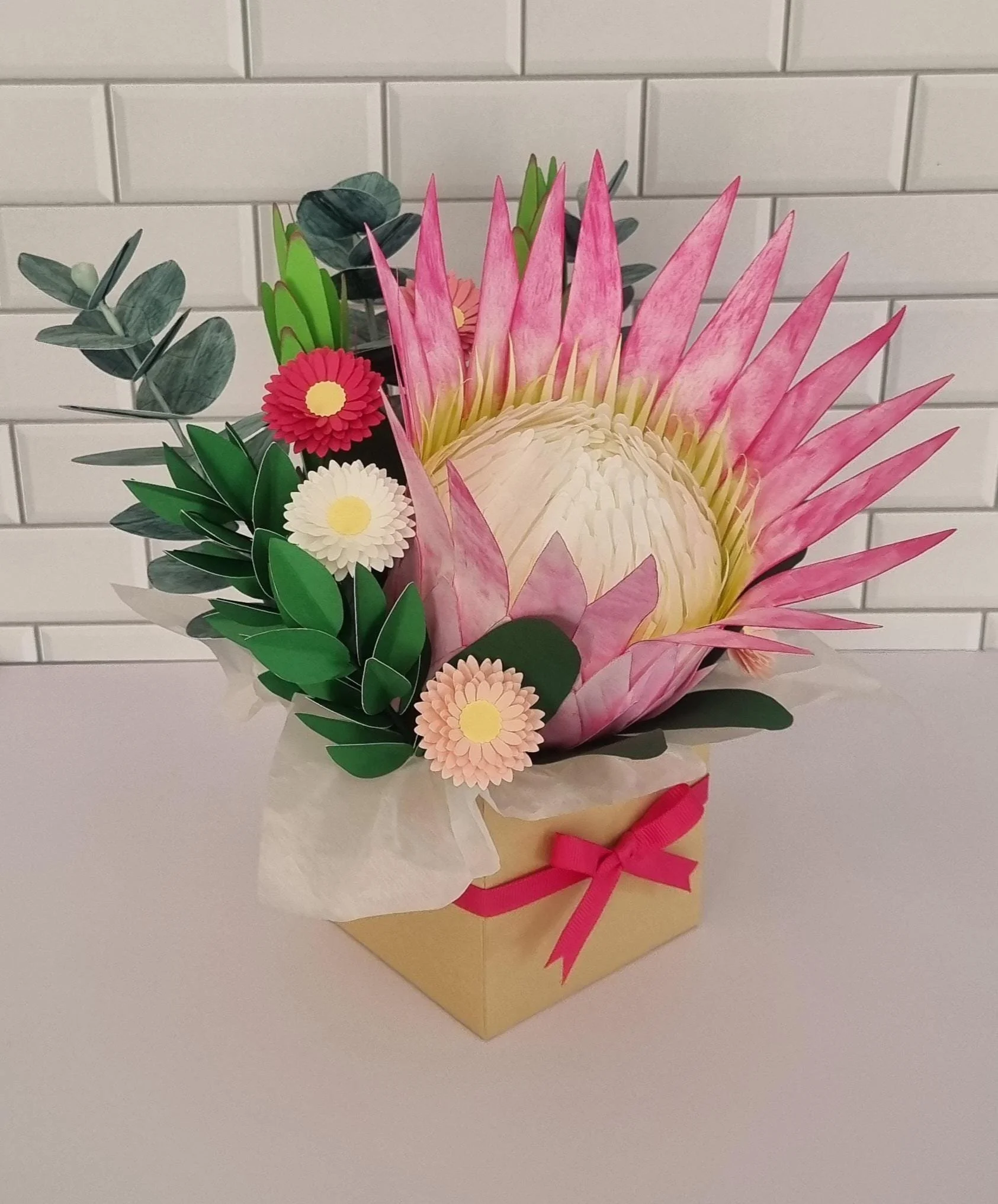 A decorative flower arrangement with pink, white, and red flowers, and green leaves in a gold box with a pink ribbon, set against a white brick wall.