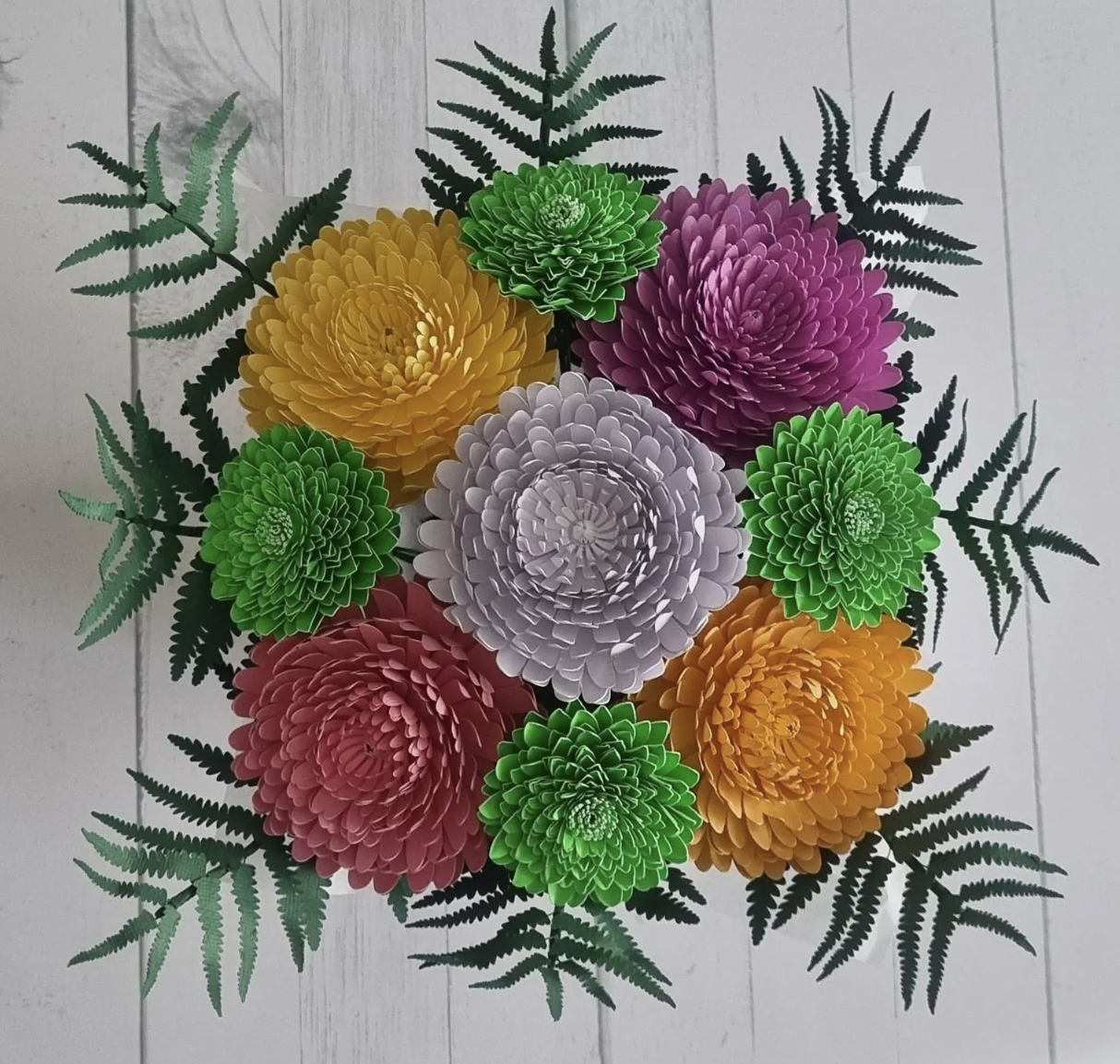 Colorful paper flower arrangement with large blooms in yellow, purple, pink, white, and green, surrounded by green paper fern leaves on white wooden background.