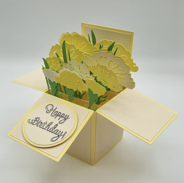 Three-dimensional handmade birthday card with yellow and green paper flowers inside a box, with a circular "Happy Birthday" message on the front.