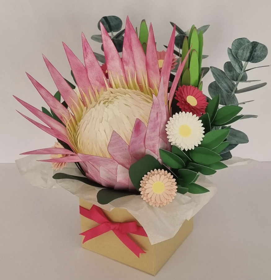 Artificial flower arrangement featuring a large pink protea, green leaves, and small pink and white daisies in a cream-colored box with a pink ribbon