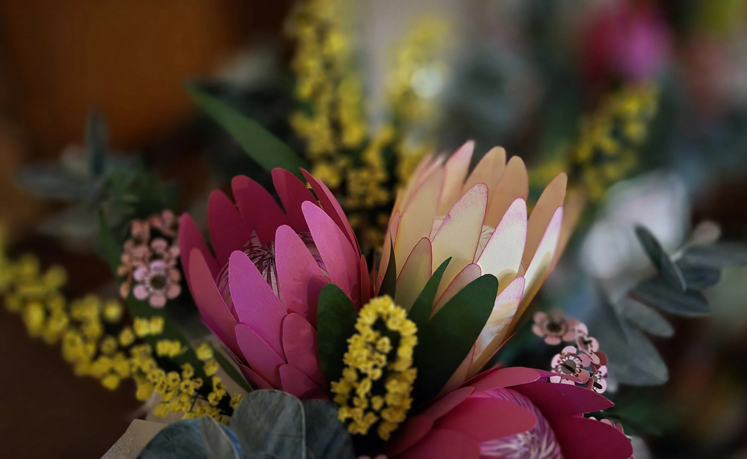 Close-up view of a colorful bouquet of native handmade paper flowers, including pink, white, and yellow blossoms, with green leaves in the background.