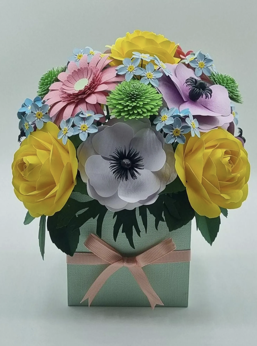 Paper flower bouquet with yellow roses, pink gerbera, white anemone, purple magnolia, blue forget-me-nots, and green chrysanthemums in a light green box with a pink ribbon.