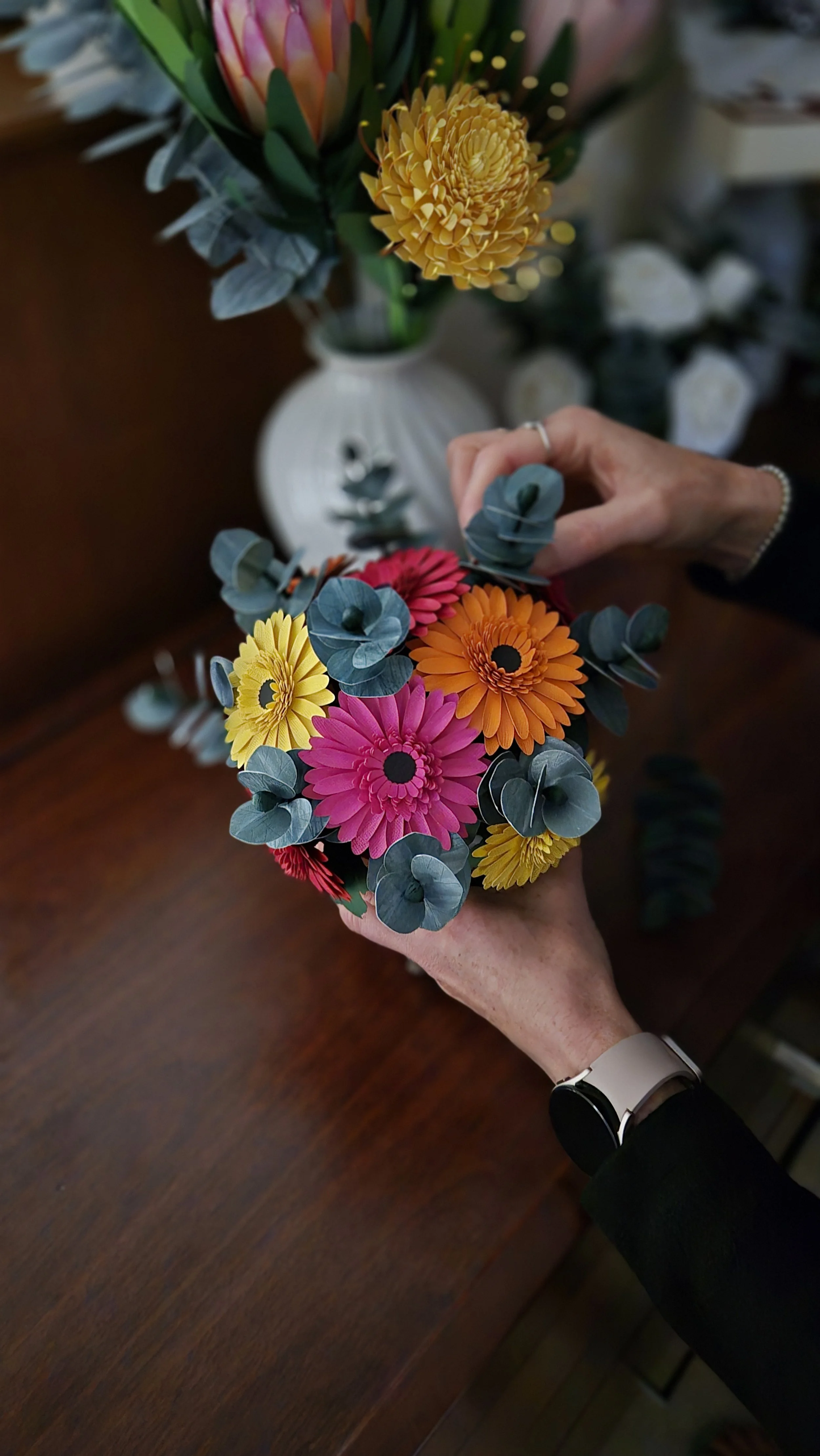 The owner Jo of Uniquely Packaged arranging a colorful bouquet of pink, orange, and yellow flowers with green leaves, with a white vase of flowers in the background on a wooden table.