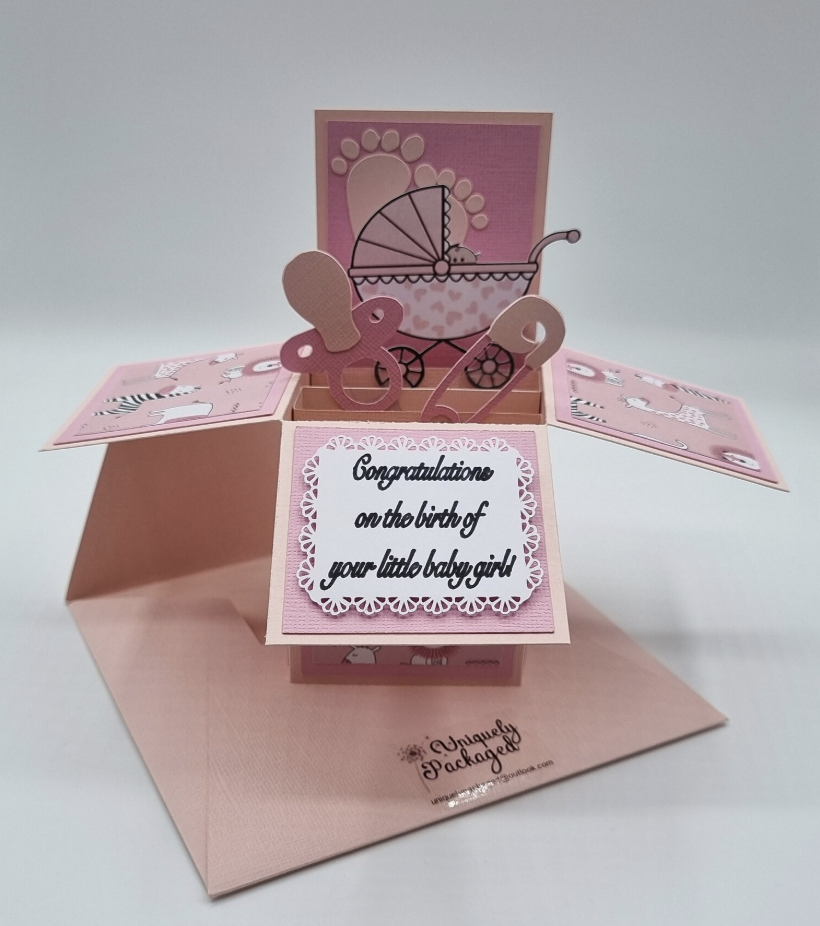 Pink pop-up greeting card with baby-themed decorations, a stroller, and a message that says "Congratulations on the birth of your little baby girl."
