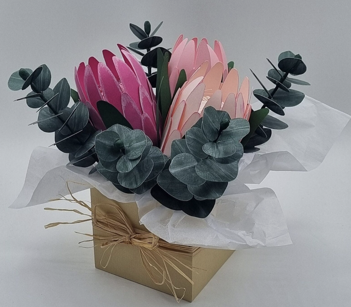Artificial flower arrangement with pink and peach petals, green eucalyptus leaves, wrapped in white tissue paper, in a beige box tied with a bow.