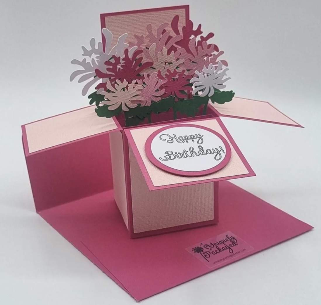 A 3D pop-up birthday card with pink tones. It features a bouquet of paper flowers and a "Happy Birthday" message on the front.