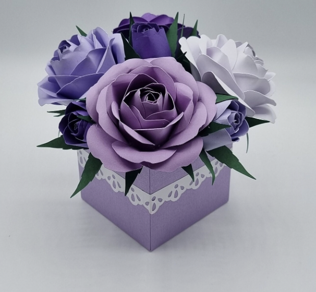 A square purple gift box with white decorative edges, filled with purple, lavender, and white paper roses and green leaves.