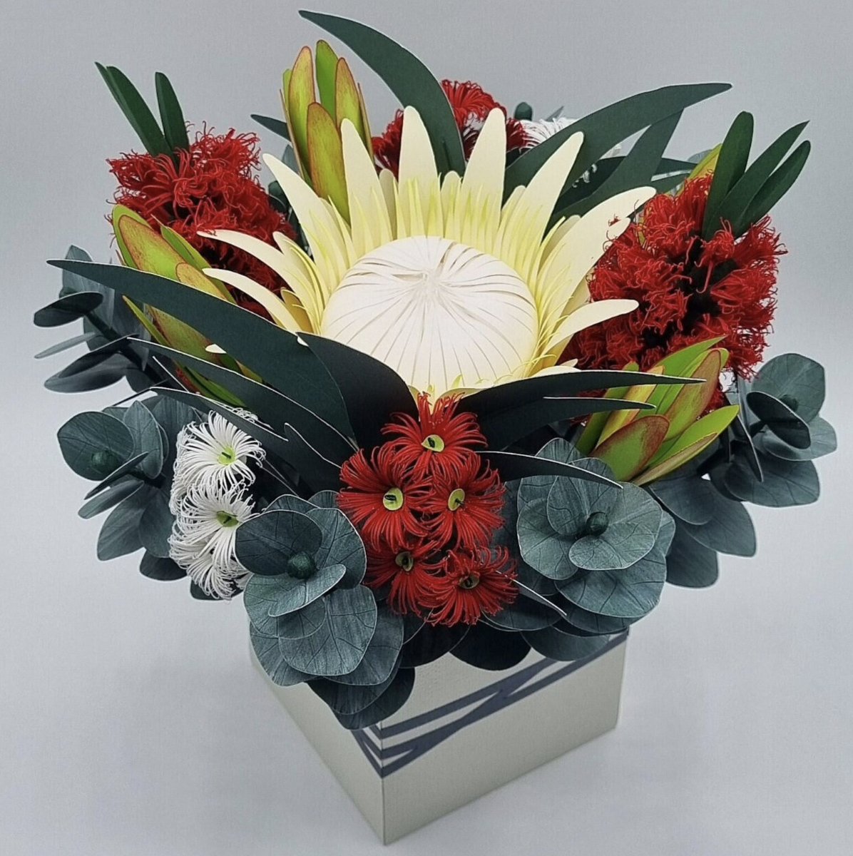 A floral arrangement in a white square vase featuring white, red, and yellow flowers with green foliage.