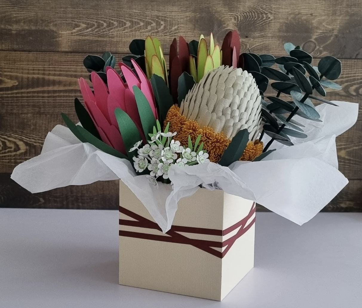 Paper flower arrangement in a cream-colored box with maroon ribbon detail, featuring pink, green, and burgundy paper flowers, cream-colored protea, orange yarn flowers, and white floral accents, set against a wood background.