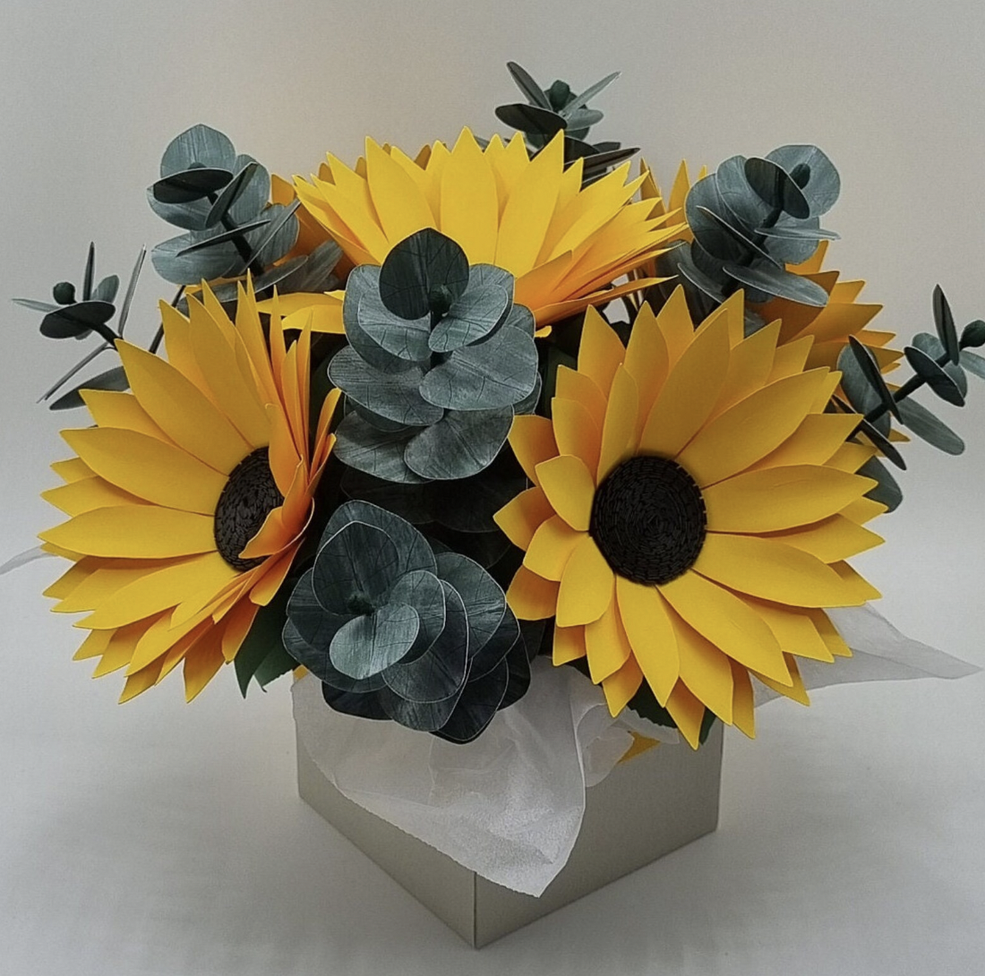 Artificial sunflower bouquet with yellow petals and dark centers, mixed with green eucalyptus leaves, in a white box with tissue paper.